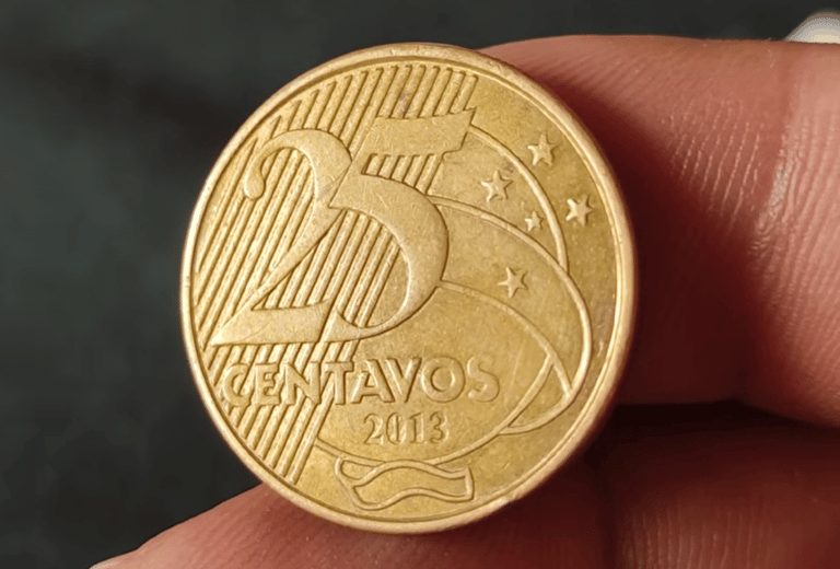 Close em detalhe de uma moeda dourada de 25 centavos de Real, do ano de 2013, sendo segurada pelos dedos de uma pessoa para análise, com foco na data e no valor.