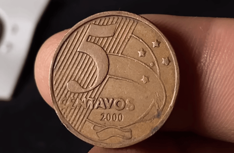 Close do reverso de uma moeda de 5 centavos do ano 2000, de cor bronze, sendo segurada por dedos. O foco está nos detalhes do cunho e na data.