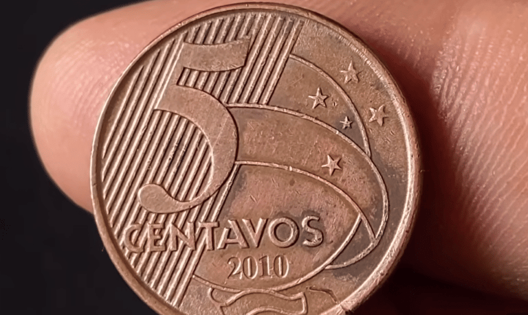 Close-up detalhado do reverso de uma moeda de 5 centavos de real do ano de 2010, sendo segurada pelos dedos de uma pessoa contra um fundo escuro.