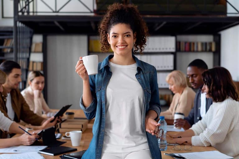 Jovens participando de uma reunião em ambiente de escritório, com foco no trabalho colaborativo, enquanto alguns estão com xícaras e outros com materiais de escritório.