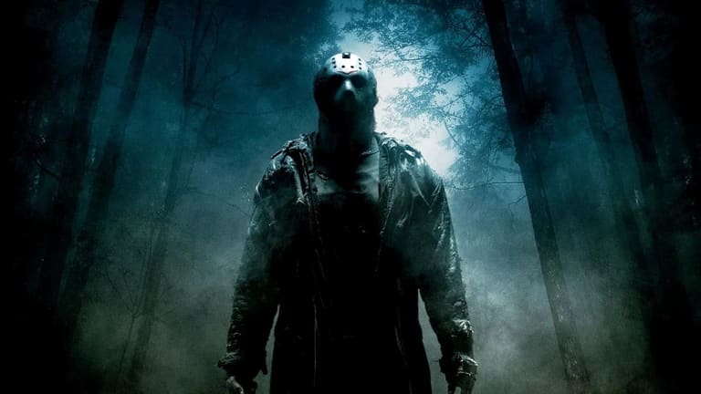 Figura sombria do personagem Jason Voorhees, de Sexta-Feira 13, usando sua icônica máscara de hóquei. Ele está em pé em uma floresta nebulosa e escura à noite, iluminada por uma luz azulada vinda de trás das árvores.