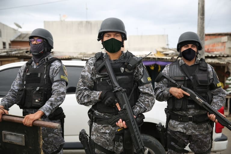 Três policiais militares do Amazonas, em uniforme tático e armados, estão em uma operação de segurança, com um cenário urbano ao fundo.