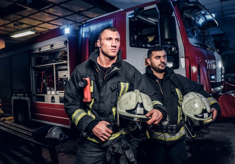 Bombeiros em frente a um caminhão de bombeiros, prontos para uma emergência, durante a noite, com equipamentos de proteção.