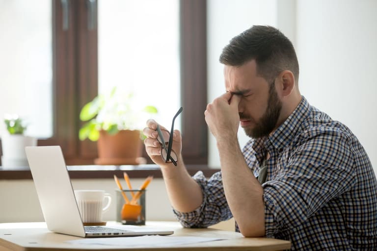 Profissional sentado à mesa de trabalho com laptop, segurando óculos e pressionando os olhos, demonstrando cansaço e sobrecarga.