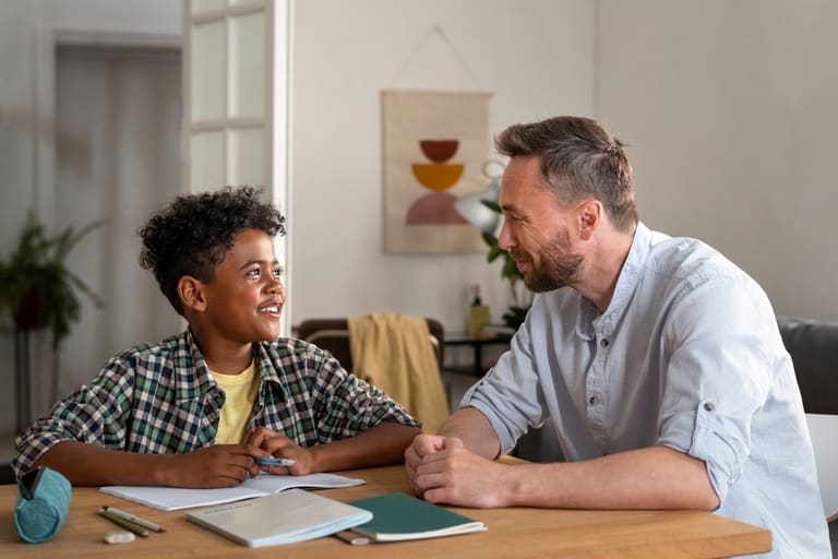 Pai e filho conversam sorridentes durante estudo em casa, com cadernos e estojo sobre a mesa