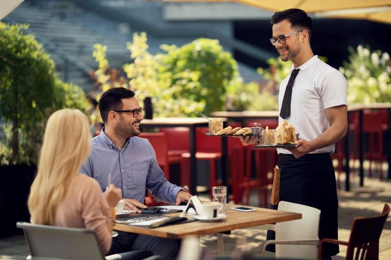 Garçom servindo bebidas e petiscos a clientes em um restaurante ao ar livre. Profissional de atendimento trabalhando com escalas 6x1.