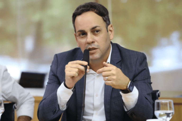 Secretário da Fazenda do Ceará de terno azul-marinho e camisa branca segura microfone durante discurso em reunião, com smartwatch no pulso