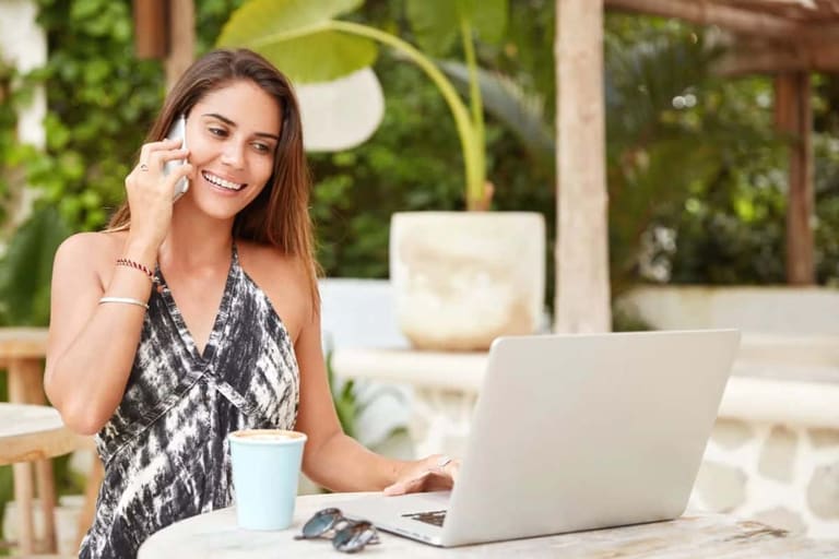Mulher sorrindo fala ao celular enquanto usa notebook em área externa, representando rotina de trabalho flexível.