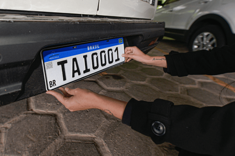Mãos posicionando placa Mercosul TAI0001 com faixa azul, QR Code e bandeira do Brasil em para-choque traseiro