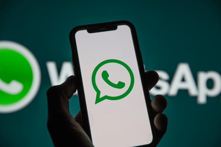 Mão segura smartphone preto com logo do WhatsApp na tela, fundo verde-azulado com marca ampliada do aplicativo
