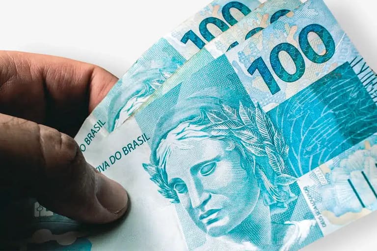 Mão segurando três cédulas de 100 reais.