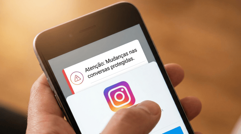 Mão segurando smartphone com notificação do Instagram sobre mudanças nas conversas protegidas e logo do app.
