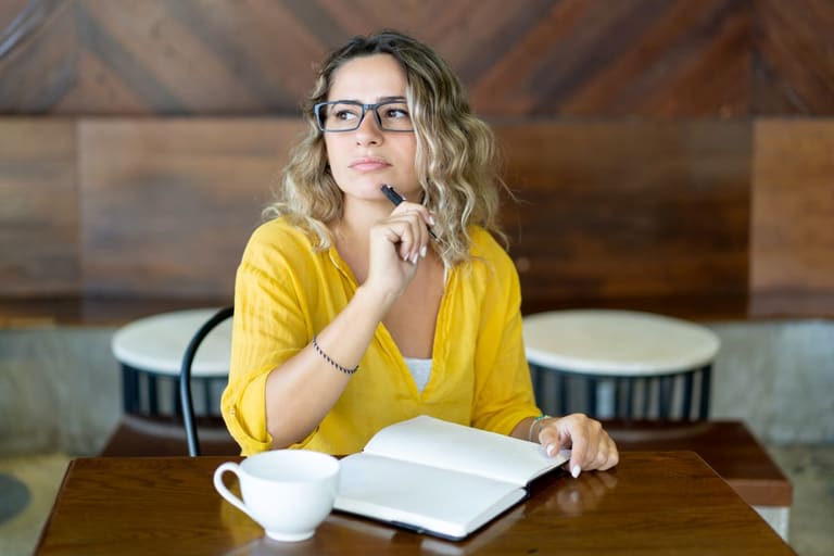 Mulher pensativa com caderno aberto em café aconchegante.