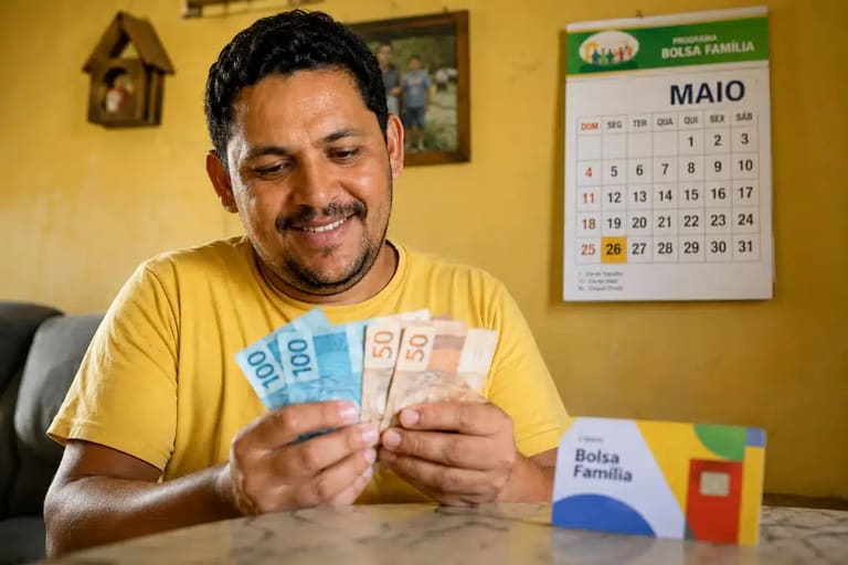 Beneficiário do Bolsa Família conta notas de reais em uma casa simples, representando o pagamento para o NIS final 7 em maio, com parcelas extras.