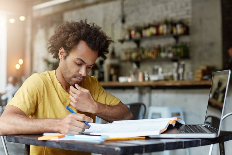 Homem de camiseta amarela estuda com livros, anotações e notebook sobre mesa de madeira em ambiente de cafeteria