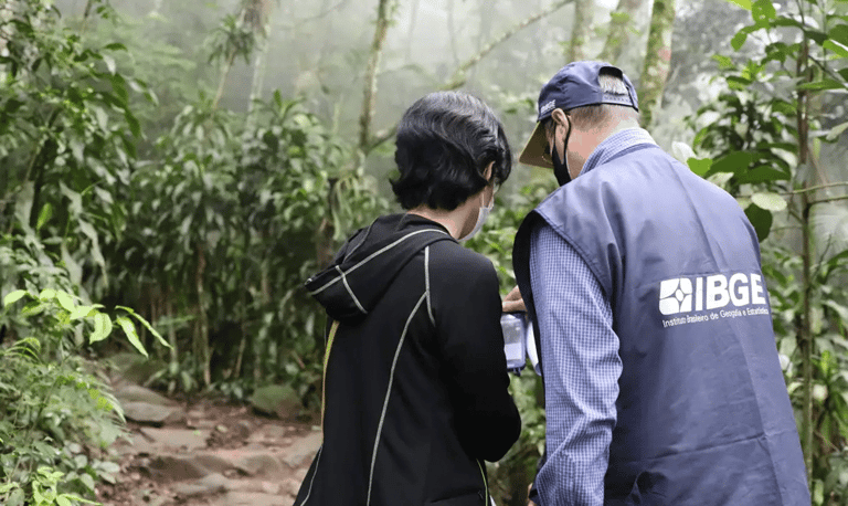 Dois servidores do IBGE com colete e boné institucionais em trabalho de campo dentro de área de mata