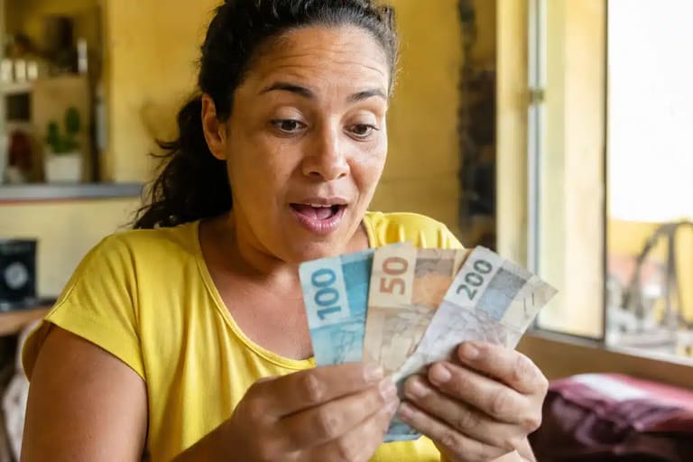Mulher brasileira surpresa segurando notas de reais, representando o pagamento único de R$ 50 mil liberado pelo Governo do Brasil.