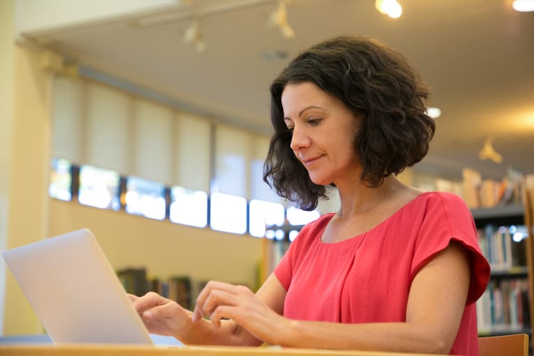 Mulher de blusa vermelha digita em notebook prateado sentada em ambiente de biblioteca com estantes de livros ao fundo
