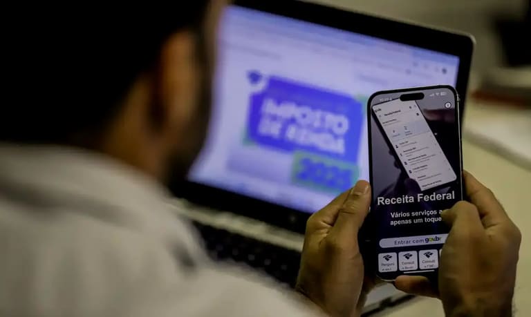 Pessoa acessando serviços da Receita Federal pelo celular enquanto declara o imposto de renda 2026 no computador.