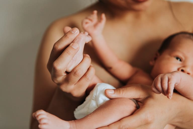 Mãe segura o bebê recém-nascido nos braços, com a mãozinha da criança apoiada em sua mão.