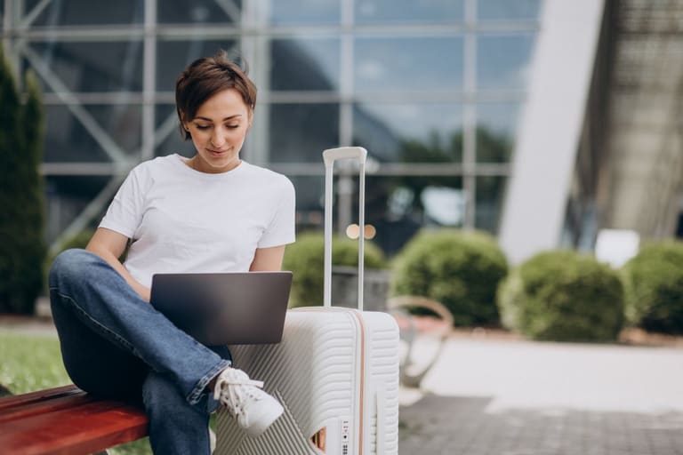 Profissional trabalhando no notebook durante viagem com mala ao lado em área externa de aeroporto.