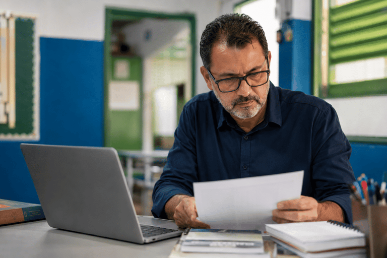 Homem de óculos e camisa azul-marinho lê documento em mesa com laptop, livros e cadernos em sala de aula.