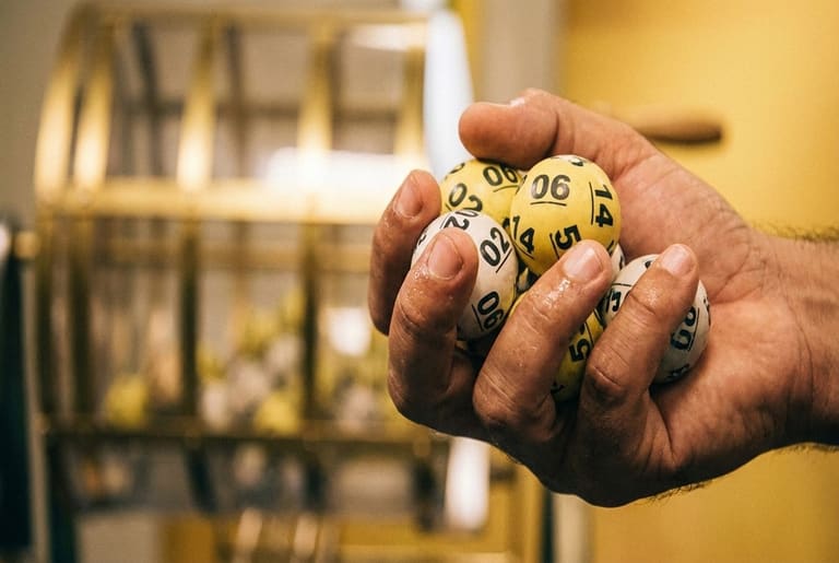 Mão masculina segurando bolas numeradas amarelas e brancas com números 02, 06, 14 e outros, globo ao fundo