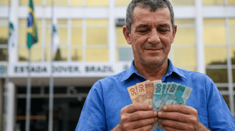 Servidor público sorri ao contar cédulas de 50 e 100 reais em frente a prédio governamental com bandeiras