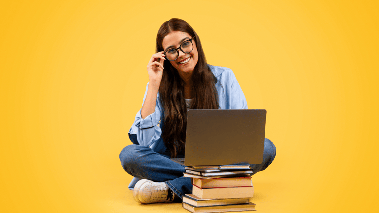 Jovem mulher de cabelo longo usando óculos e sorrindo, sentada com notebook sobre pilha de livros em fundo amarelo.