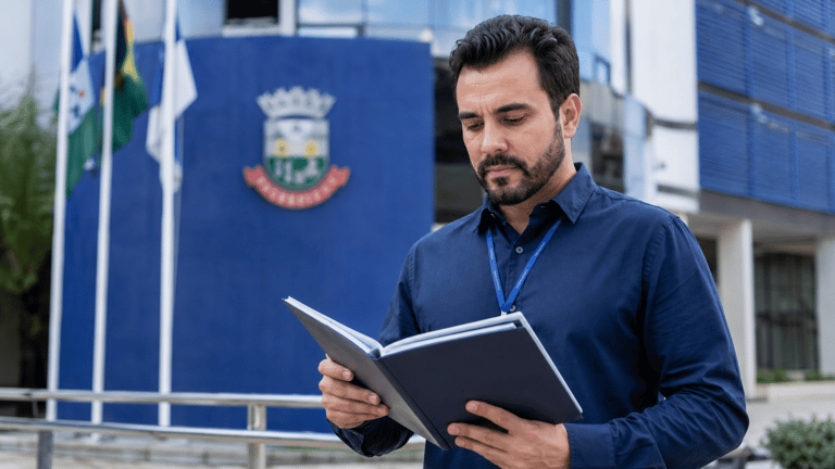 Homem de barba e crachá azul lendo pasta preta diante de prédio institucional com bandeiras.