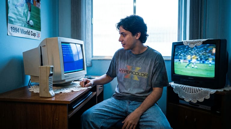 Jovem usa computador antigo de tubo ao lado de TV de tubo exibindo futebol